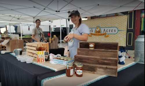 A vendor at a market displays honey jars and products, promoting a honey farm with a cheerful smile.