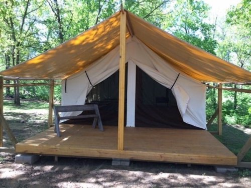 A glamping tent with a wooden deck and a brown canopy, set in a green outdoor area surrounded by trees.