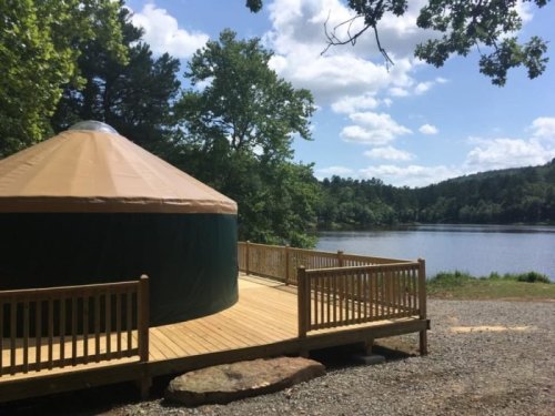 A yurt on a wooden deck by a serene lake, surrounded by trees and under a partly cloudy sky.