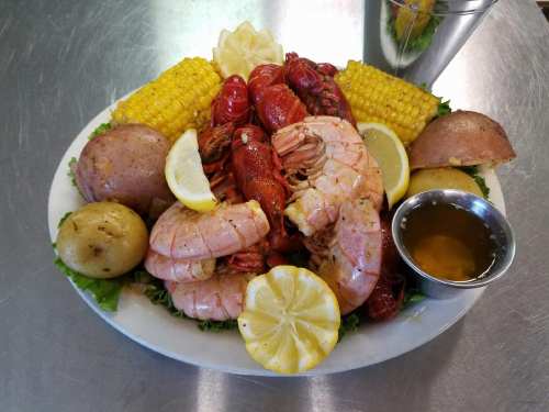 A seafood platter featuring shrimp, crawfish, corn, potatoes, and lemon wedges, served with a side of dipping sauce.