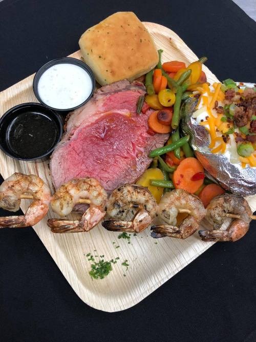 A wooden platter with prime rib, shrimp, baked potato, mixed vegetables, a roll, and dipping sauces.