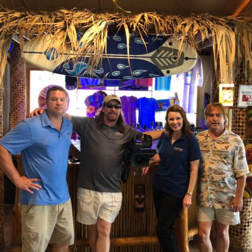 Four people pose together in a tropical-themed setting with surfboards and colorful decor in the background.