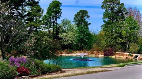 A serene pond surrounded by lush greenery and colorful flowers, featuring a fountain in the center under a clear blue sky.