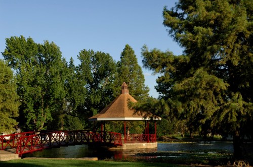 A red bridge leads to a gazebo surrounded by lush green trees and a calm waterway under a clear blue sky.