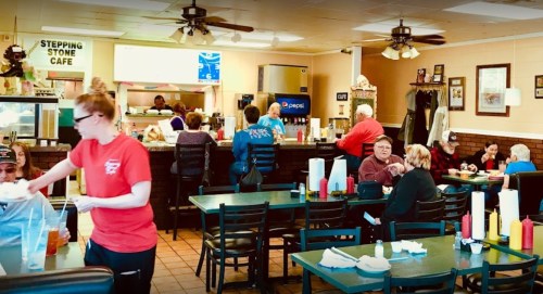 A busy café interior with patrons dining, a server in red, and a counter with staff preparing food.