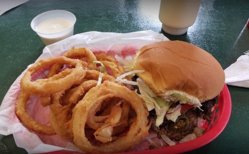 A basket with a burger topped with lettuce and pickles, accompanied by crispy onion rings and a small cup of sauce.