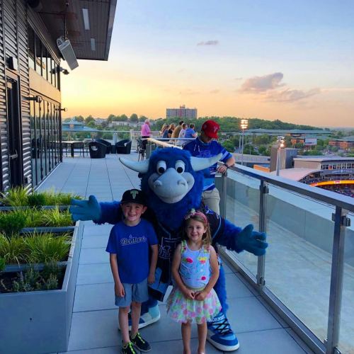 A blue mascot poses with two children on a balcony during sunset, with a crowd and cityscape in the background.