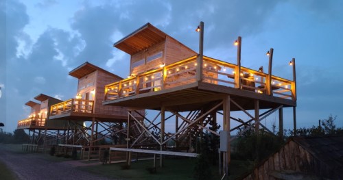 Three elevated wooden cabins with warm lights, set against a twilight sky, surrounded by greenery.