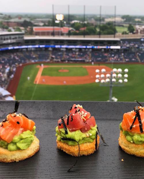 Three colorful appetizers topped with salmon and avocado, with a baseball field and crowd in the background.