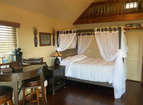Cozy bedroom with a four-poster bed, wooden accents, and a small dining area. Natural light from a window.