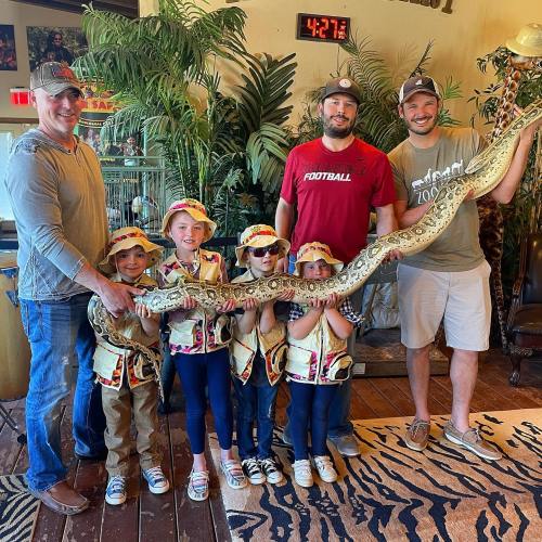 A group of four adults and five children hold a large snake in a tropical-themed setting.
