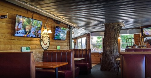 Cozy restaurant interior with wooden walls, booths, and TVs, featuring a tree trunk in the center.