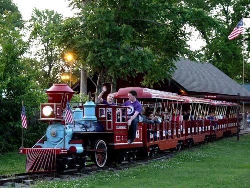 A colorful miniature train with passengers rides through a park, surrounded by trees and American flags.