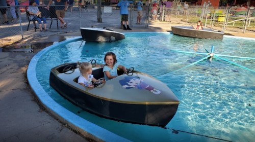 Two children in small boats on a water ride, smiling and enjoying a sunny day at an amusement park.