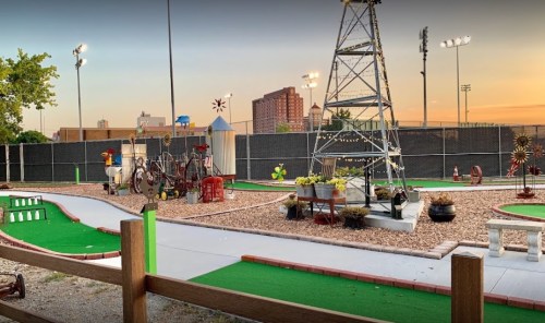 A mini-golf course with decorative windmills and a tower, set against a sunset skyline and city buildings in the background.