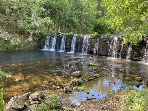 A serene waterfall cascades over rocks into a calm pool, surrounded by lush greenery and trees.