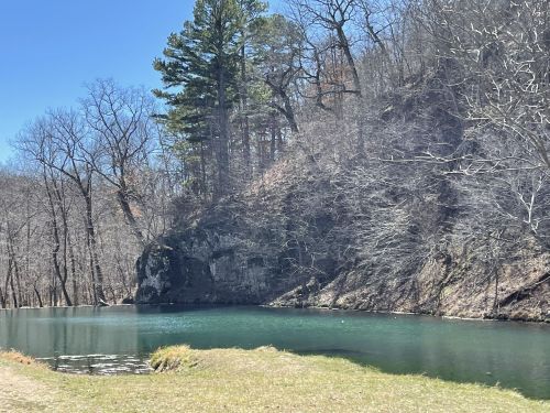 A serene pond surrounded by bare trees and a rocky hillside under a clear blue sky.