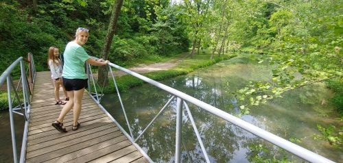 A woman and a girl stand on a wooden bridge over a calm, reflective stream surrounded by lush greenery.