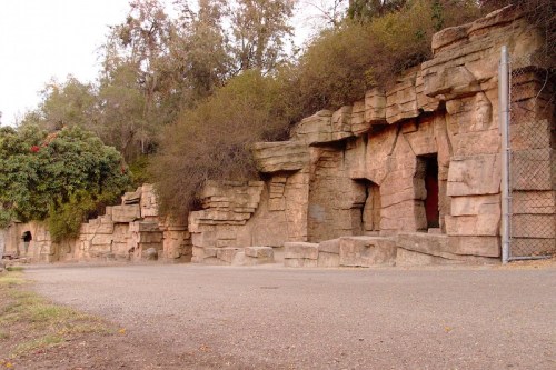 Rocky cliffside with natural stone formations and sparse vegetation, alongside a gravel path and a fence.