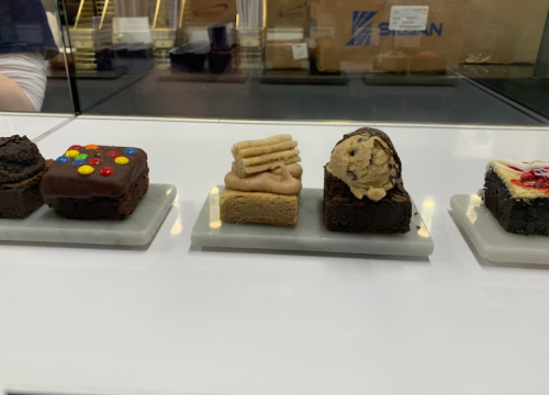 A display of assorted brownies topped with colorful candies, cream, cookies, and fruit on small white plates.