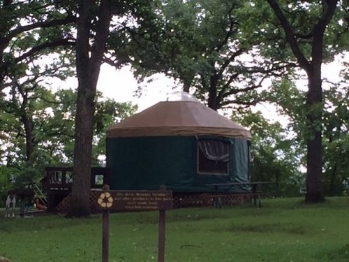 A green yurt with a brown roof sits in a grassy area surrounded by trees, with a recycling sign in the foreground.