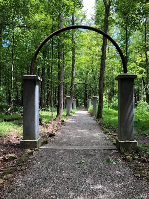 A serene forest path lined with archways, surrounded by lush green trees and dappled sunlight.