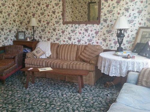Cozy living room with floral wallpaper, a striped sofa, a lace tablecloth, and vintage decor.