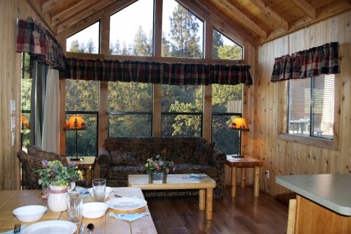 Cozy wooden cabin interior with large windows, a sofa, and a dining table set for a meal.