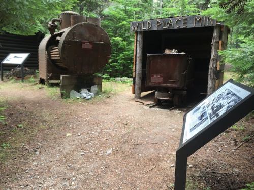Historic mining equipment displayed in a forested area, featuring a large machine and a wooden structure labeled "Wild Place Mine."