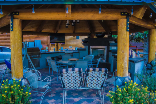 A cozy outdoor kitchen with a grill, seating area, and stone fireplace, surrounded by flowers and wooden beams.