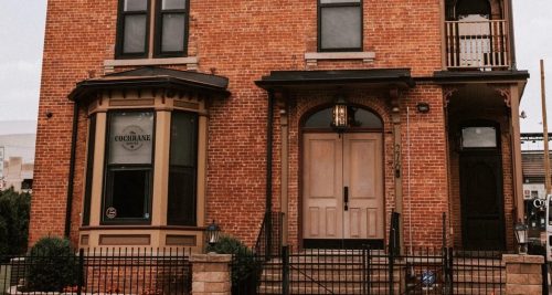 A brick building with large wooden doors and decorative windows, featuring a fenced front yard.