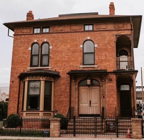 A large, red brick building with arched windows and a decorative porch, surrounded by a black fence.