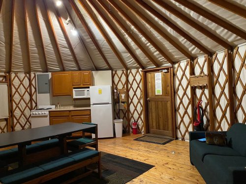 Interior of a yurt featuring wooden walls, a kitchen area, dining table, and a door leading outside.