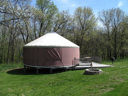 A yurt with a wooden deck sits in a grassy area surrounded by trees under a clear blue sky.