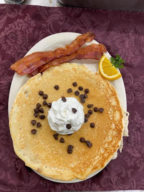 A large pancake topped with whipped cream and chocolate chips, served with bacon and an orange slice on a plate.