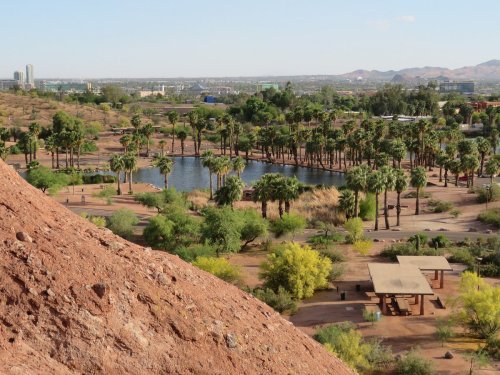 A scenic view of a park with palm trees, a pond, and mountains in the background, seen from a rocky elevation.
