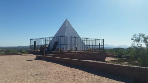 A white pyramid structure surrounded by a fence, set against a clear blue sky and desert landscape.