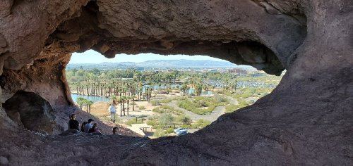View through a rock formation, showcasing a lush landscape with palm trees and a lake in the distance.
