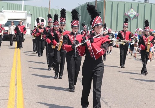 A marching band in red and black uniforms parades down a street, playing instruments and wearing feathered hats.