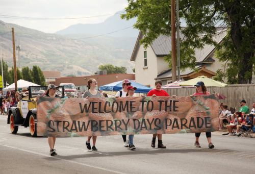 A group of people carrying a large banner that reads "Welcome to the Strawberry Days Parade" during a festive parade.