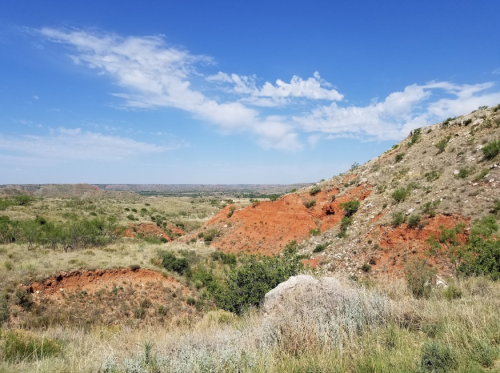 A scenic view of rolling hills with red soil under a bright blue sky and scattered clouds.