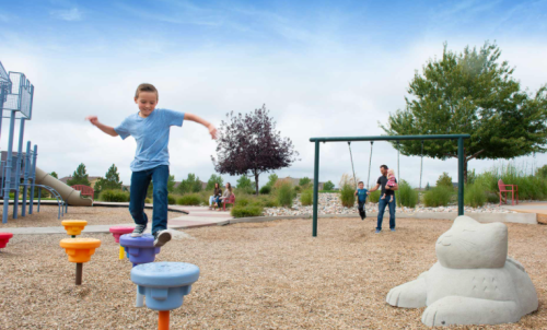 A child balances on colorful stepping stones in a playground, with swings and a slide in the background.