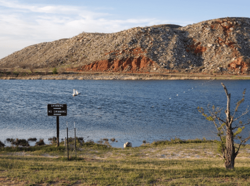 A serene lake with a "No Swimming" sign, surrounded by rocky hills and a small sailboat on the water.
