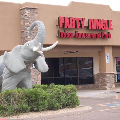 A large elephant statue stands outside the "Party Jungle" indoor amusement park, with a stone building in the background.