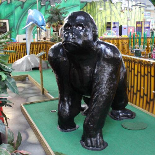 A large black gorilla statue sits on a mini-golf course surrounded by colorful decor and plants.