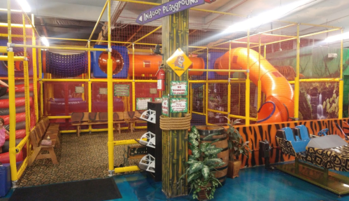 Colorful indoor playground with slides, climbing structures, and seating areas, surrounded by safety netting.
