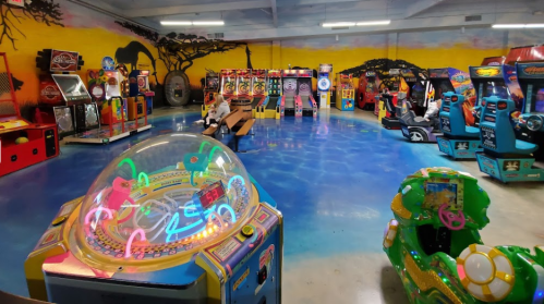 A colorful arcade filled with various games, including racing and shooting machines, under a vibrant mural.