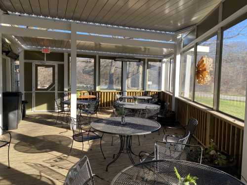 A sunlit screened porch with metal tables and chairs, surrounded by windows and wooden walls.