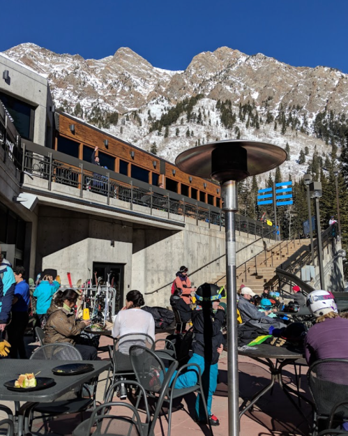 A busy outdoor ski area with people dining and enjoying the sun, surrounded by snow-covered mountains.