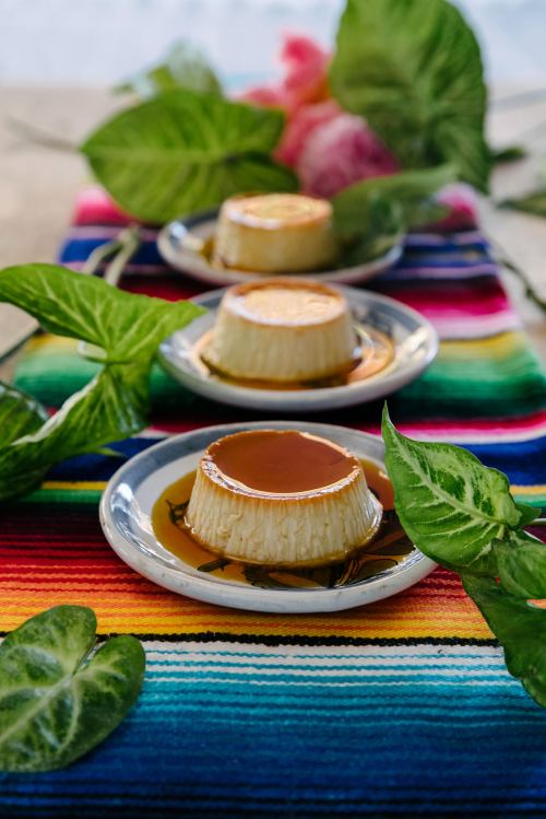 Three flans on white plates, drizzled with caramel, arranged on a colorful striped tablecloth with green leaves.